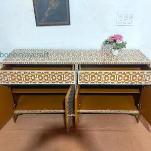 May include: A mustard-coloured cabinet with two drawers and two doors, featuring a white geometric inlay pattern. The cabinet doors are open, revealing interior shelves. The top of the cabinet has a floral arrangement. The text "ikboneinlaycraft" is visible on the top left.