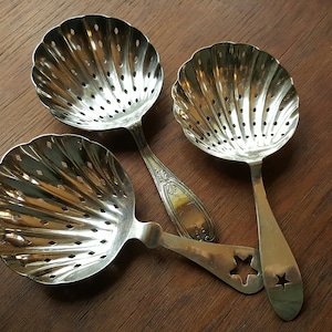 May include: Three silver serving spoons with shell-shaped bowls and pierced designs. One spoon has a handle with a star cutout. The spoons are arranged on a dark wooden surface.