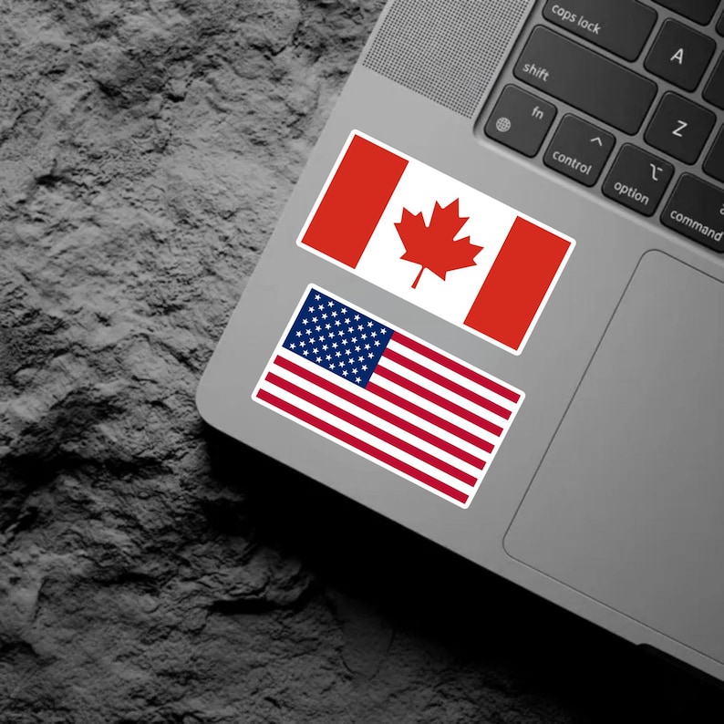 May include: Two rectangular stickers applied to the surface of a laptop. The top sticker features the red and white Canadian flag with a central red maple leaf. The bottom sticker displays the red, white, and blue flag of the United States.
