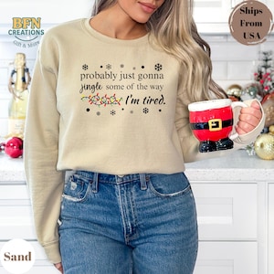 May include: A sand-colored sweatshirt with the text "probably just gonna jingle some of the way I'm tired." The design includes snowflakes and Christmas lights. A red and white Santa mug is held in the hand.