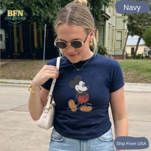May include: Navy blue t-shirt featuring a classic Mickey Mouse graphic. The cartoon character is depicted in his iconic red shorts, brown shoes, and white gloves. The shirt is a solid navy color.