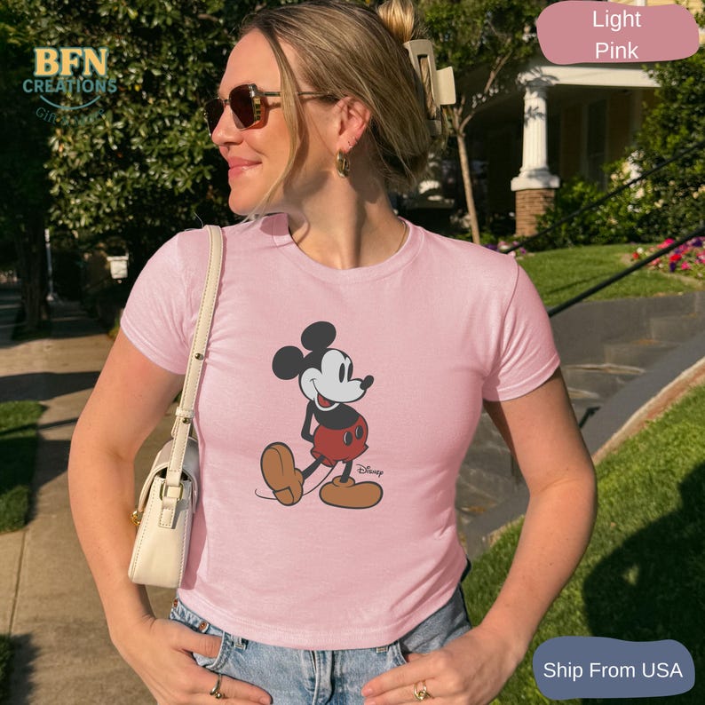 May include: Light pink t-shirt featuring a classic Mickey Mouse graphic. The cartoon character is depicted in his iconic pose, wearing red shorts, brown shoes, and white gloves. The shirt is a light pink color.