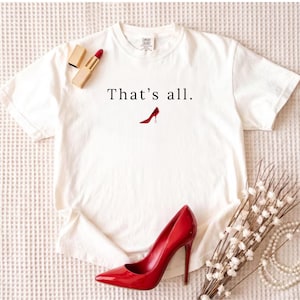 May include: White t-shirt with the text "That's all." and a red high heel graphic. A red high heel shoe and lipstick are next to the shirt. The shirt is made of a soft material.