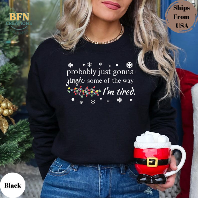 May include: Black sweatshirt with the text "probably just gonna jingle some of the way I'm tired." The design includes snowflakes and a string of Christmas lights. A Santa mug is held in the hand.