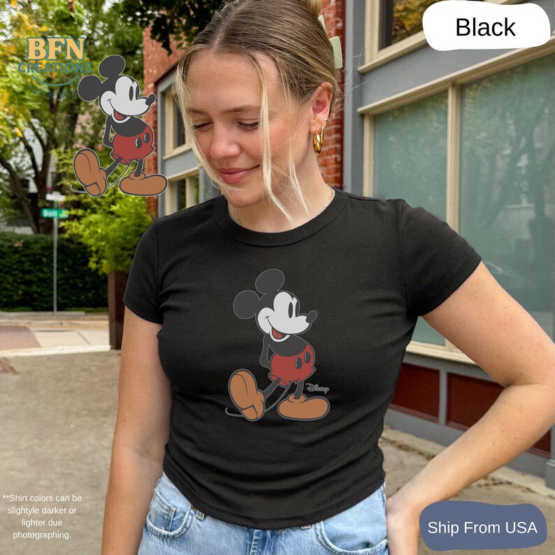 May include: Black t-shirt featuring a graphic of Mickey Mouse in a classic pose. The cartoon character is depicted in his signature red shorts, white gloves, and brown shoes. The shirt is a crew neck style.