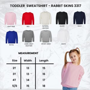 May include: A light pink toddler sweatshirt from Rabbit Skins, with a measurement chart showing sizes 2T to 5/6. The image displays various colors, including red, black, navy, royal blue, and gray. The text "TODDLER SWEATSHIRT - RABBIT SKINS 3317" is at the top.