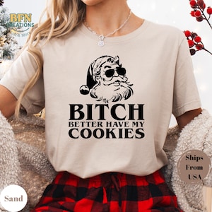 May include: Sand-colored t-shirt featuring a black graphic of Santa Claus wearing sunglasses, with the text "BITCH BETTER HAVE MY COOKIES" in bold letters. The shirt is paired with red and black plaid pajama pants.