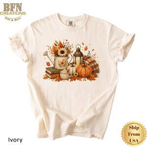 May include: An ivory t-shirt featuring a graphic print of a fall-themed arrangement. The design includes a watering can filled with sunflowers, a lantern, stacked books, a mug, pumpkins, and a plaid blanket surrounded by autumn leaves.