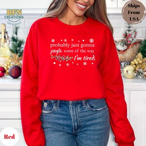 May include: A red crewneck sweatshirt with the text "probably just gonna jingle some of the way I'm tired." The design includes snowflakes and a string of Christmas lights. The sweatshirt is a solid red color.