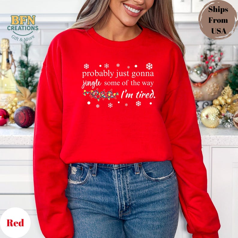 May include: A red crewneck sweatshirt with the text "probably just gonna jingle some of the way I'm tired." The design includes snowflakes and a string of Christmas lights. The sweatshirt is a solid red color.