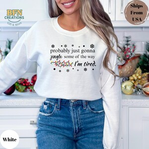 May include: White sweatshirt with the text "probably just gonna jingle some of the way I'm tired." The design includes snowflakes and a string of Christmas lights. The sweatshirt is a casual, festive garment.