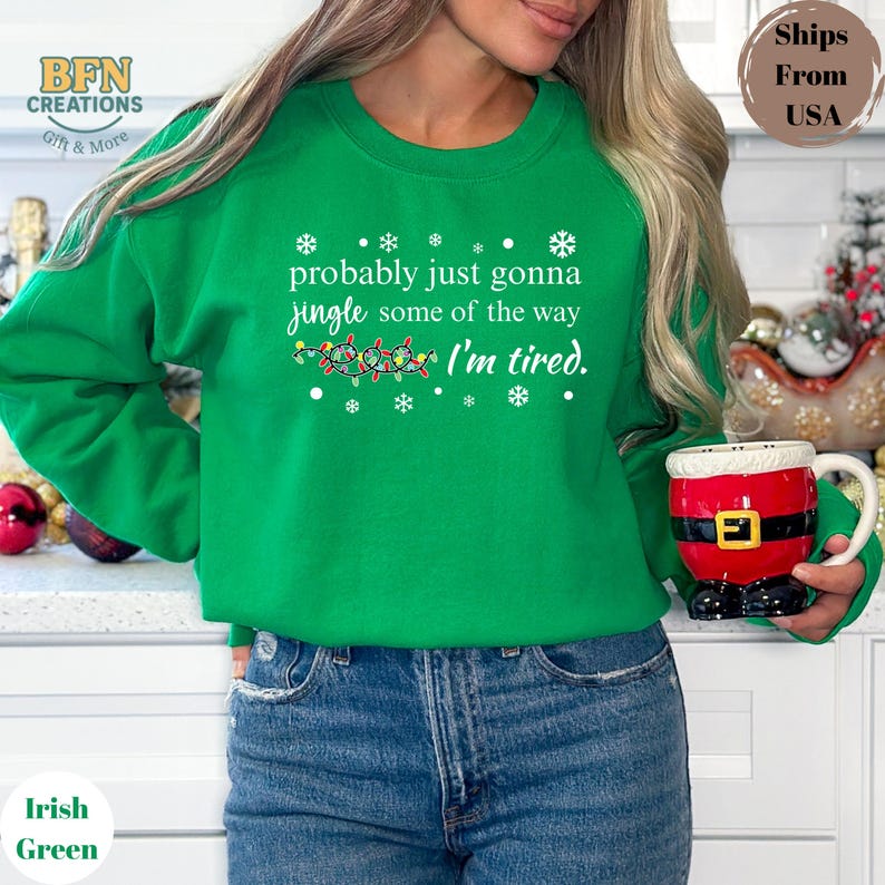 May include: Irish green sweatshirt with the text "probably just gonna jingle some of the way, I'm tired." The design includes snowflakes and a string of Christmas lights. A festive mug with a Santa Claus design is held in the hand.