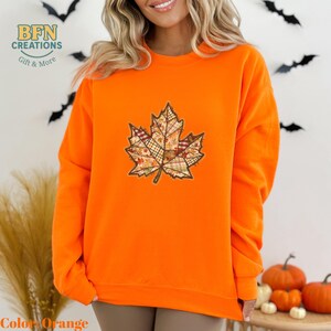 May include: Bright orange sweatshirt featuring a detailed leaf design. The leaf is composed of various fabric patterns in autumn colors. The sweatshirt has long sleeves and a crew neck. The text "BFN CREATIONS Gift & More" is visible in the upper left corner.