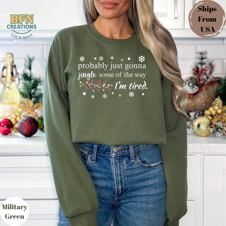 May include: Military green sweatshirt with the text "probably just gonna jingle some of the way I'm tired." The design includes snowflakes and a string of Christmas lights. The sweatshirt is a casual, festive garment.