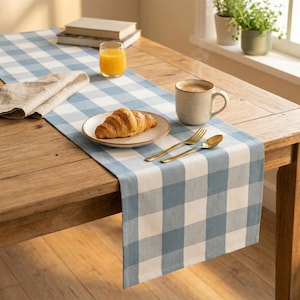 Puede incluir: Una mesa de madera está preparada para el desayuno con un camino de mesa a cuadros azul y blanco. Se ve un plato con un cruasán, una taza de café, un vaso de zumo de naranja y cubiertos dorados.