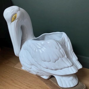 May include: A white ceramic pelican planter with a glossy finish. The bird has a long beak, a curved neck, and detailed feathering on its wings and body. The eye is a contrasting gold colour. The planter sits on a wooden base.