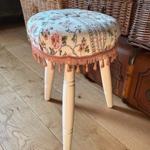 May include: A round stool with three white tapered wooden legs and a padded seat upholstered in a cream fabric featuring a floral pattern in shades of pink, green, and yellow. The edge of the seat is finished with a decorative pink fringe trim.