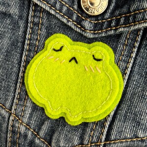 Peut inclure: Un écusson en feutre vert citron en forme de grenouille avec un contour et des traits du visage cousus. La grenouille a les yeux fermés, un petit sourire et des marques de blush. L'écusson est fixé à une veste en jean.