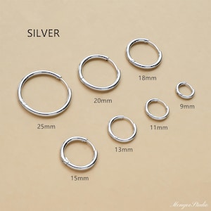 Puede incluir: Pendientes de aro plateados en varios tamaños: 9mm, 11mm, 13mm, 15mm, 18mm, 20mm y 25mm. La palabra "SILVER" está impresa en la parte superior. Los pendientes se muestran sobre una superficie marrón claro.