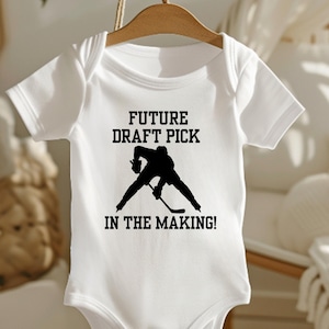 Puede incluir: Body de bebé blanco con el texto negro "FUTURE DRAFT PICK IN THE MAKING!". Una silueta negra de un jugador de hockey aparece debajo del texto. El body está colgado de una percha de madera.