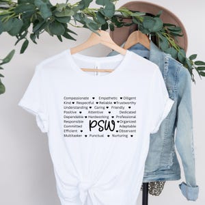 Può includere: Maglietta bianca con la scritta "PSW" e un elenco di attributi positivi come "compassionevole" e "affidabile". La maglietta è appesa a una gruccia di legno, con una giacca di jeans e un cappello sullo sfondo.