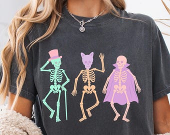 May include: Dark gray t-shirt featuring a colorful skeleton graphic. The skeletons are dressed in a top hat, cat ears, and a cape. The graphic is in pastel colors, perfect for Halloween or spooky season.