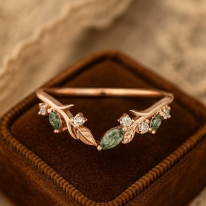 May include: A rose gold ring with a leaf and gemstone design. The ring features small green gemstones and clear accent stones, set in a delicate leaf pattern. The ring is displayed on a brown velvet surface.