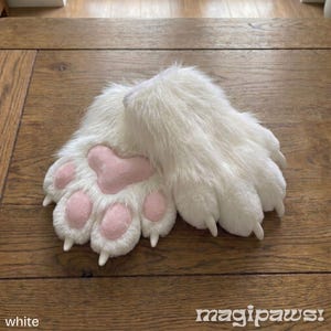 Puede incluir: Un par de guantes blancos y peludos con almohadillas rosas y garras. Los guantes est&aacute;n hechos de un material suave y afelpado y est&aacute;n dise&ntilde;ados para parecerse a las patas de los animales. El texto "magipaws!" es visible.