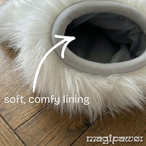 Puede incluir: Primer plano de una cama para mascotas con un exterior blanco y esponjoso y un forro gris, suave y c&oacute;modo. La cama tiene una abertura circular y est&aacute; colocada sobre una superficie de madera. El texto "soft, comfy lining" es visible.
