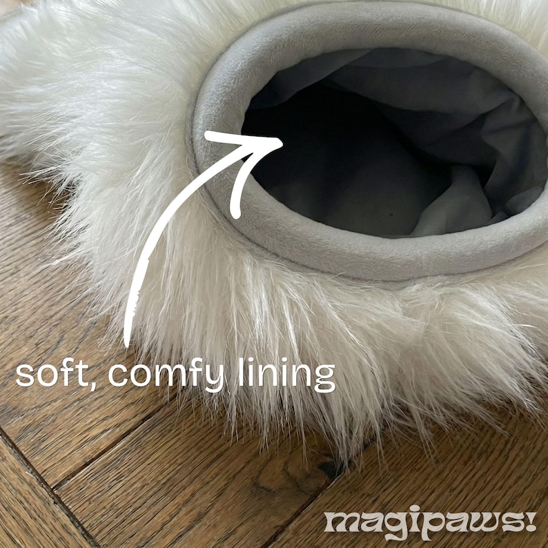 Puede incluir: Primer plano de una cama para mascotas con un exterior blanco y esponjoso y un forro gris, suave y c&oacute;modo. La cama tiene una abertura circular y est&aacute; colocada sobre una superficie de madera. El texto "soft, comfy lining" es visible.