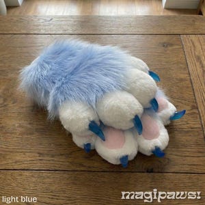 Puede incluir: Una pata de peluche azul claro con almohadillas blancas y garras azules. La pata tiene una textura esponjosa y est&aacute; dise&ntilde;ada para parecerse a la pata de un animal. El texto "magipaws!" est&aacute; en la esquina inferior derecha.