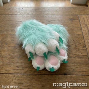 Puede incluir: Guantes de pata de piel sint&eacute;tica verde claro con almohadillas blancas y detalles en rosa. Los guantes tienen garras verdes y est&aacute;n etiquetados como "magipaws!". El texto "light green" tambi&eacute;n es visible.