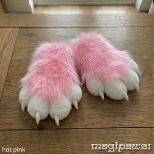 Puede incluir: Un par de zapatillas de pata peludas rosas y blancas. Las zapatillas tienen un aspecto afelpado y texturizado con garras blancas. El texto "magipaws!" est&aacute; en la esquina inferior derecha y "hot pink" en la inferior izquierda.