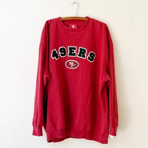May include: A red crewneck sweatshirt with the word "49ERS" in white and black lettering across the chest. The sweatshirt has long sleeves and a small logo patch near the neckline. The garment is hanging on a wooden hanger.