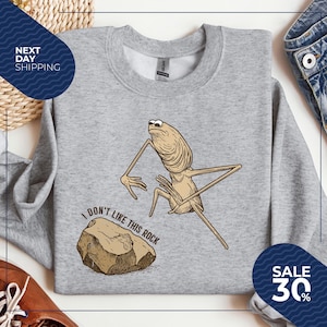 May include: A light gray sweatshirt featuring a cartoon character with long limbs and the text "I DON'T LIKE THIS ROCK." The character is next to a brown rock. A blue circle with "SALE 30%" is on the bottom right.