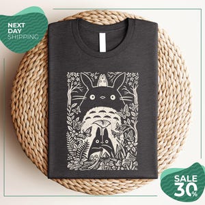 a gray t-shirt with a black and white graphic design depicting a cat surrounded by various natural elements, such as flowers, leaves, and mushrooms. The t-shirt is placed on a woven placemat, and there are two green circular signs in the image. One sign advertises "Next Day Shipping" and the other sign advertises a "SALE 30%".