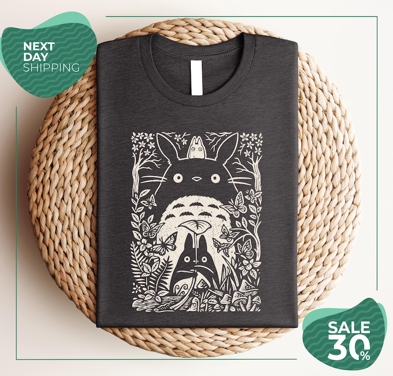 a gray t-shirt with a black and white graphic design depicting a cat surrounded by various natural elements, such as flowers, leaves, and mushrooms. The t-shirt is placed on a woven placemat, and there are two green circular signs in the image. One sign advertises "Next Day Shipping" and the other sign advertises a "SALE 30%".