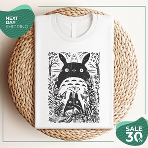 a white t-shirt with a black and white graphic design depicting a cat surrounded by various plants and flowers. The t-shirt is placed on a woven placemat, and there are two green circular sale signs in the image.
