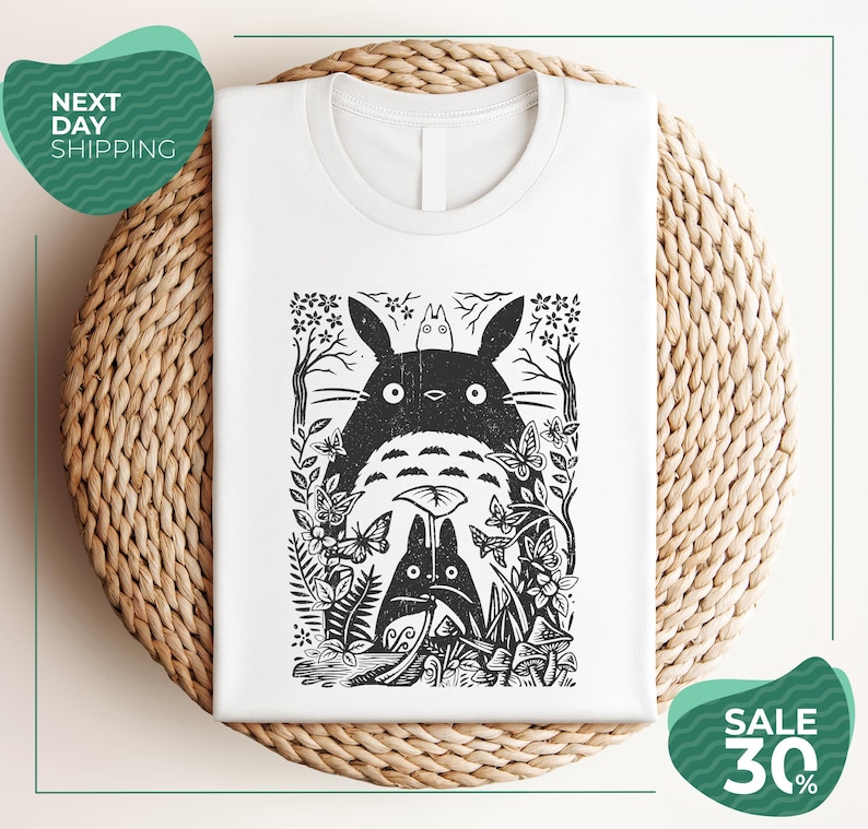 a white t-shirt with a black and white graphic design depicting a cat surrounded by various plants and flowers. The t-shirt is placed on a woven placemat, and there are two green circular sale signs in the image.
