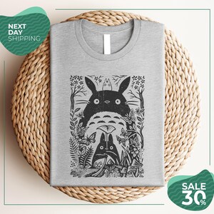 a gray t-shirt with a black and white graphic design depicting a cat surrounded by various plants and flowers. The t-shirt is placed on a woven placemat, and there are two green circular sale signs in the image.