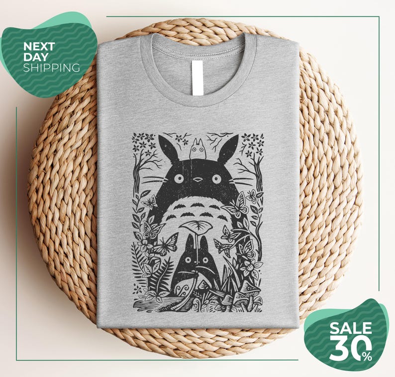 a gray t-shirt with a black and white graphic design depicting a cat surrounded by various plants and flowers. The t-shirt is placed on a woven placemat, and there are two green circular sale signs in the image.