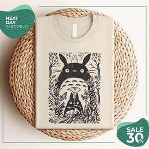 a beige t-shirt with a black and white graphic design depicting a cat surrounded by various plants and flowers. The t-shirt is placed on a woven basket, and there are two green circular signs in the image. One sign reads "Next Day Shipping" and the other sign says "SALE 30%".
