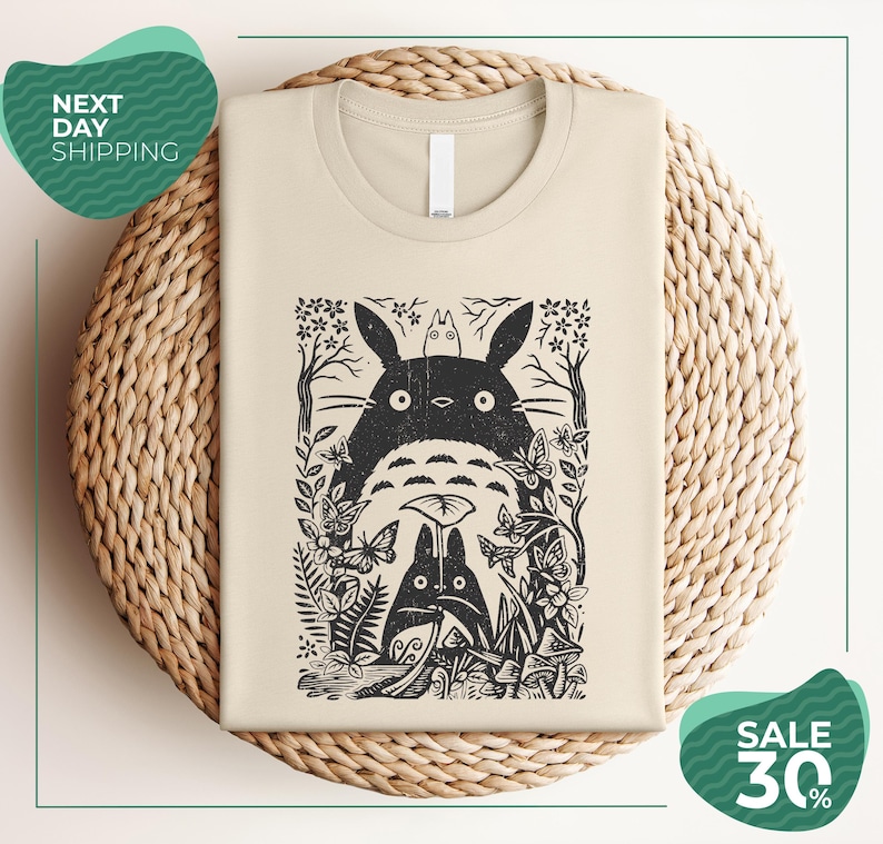 a beige t-shirt with a black and white graphic design depicting a cat surrounded by various plants and flowers. The t-shirt is placed on a woven basket, and there are two green circular signs in the image. One sign reads "Next Day Shipping" and the other sign says "SALE 30%".