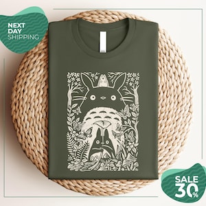 a green t-shirt with a white graphic design depicting a cat surrounded by various natural elements, such as leaves, flowers, and mushrooms. The t-shirt is displayed on a woven placemat, and there are two green circular signs in the image, one indicating "Next Day Shipping" and the other promoting a "SALE 30%".