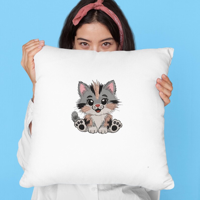 May include: White square pillow featuring an embroidered cartoon kitten. The kitten has gray, brown, and black fur, with pink ears and a smiling face. The pillow is held up against a light blue background.