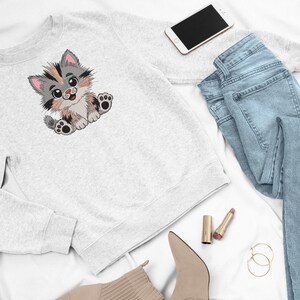 May include: A light gray sweatshirt featuring an embroidered cartoon kitten with gray, black, and tan fur. The kitten is sitting with its paws out. The image also includes light blue jeans, a smartphone, lipstick, earrings, and beige boots.