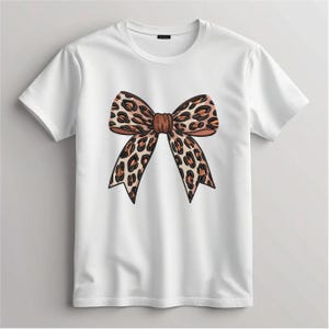May include: White t-shirt featuring a large, detailed bow design. The bow has a leopard print pattern in shades of brown and tan. The shirt is a classic crew neck style.