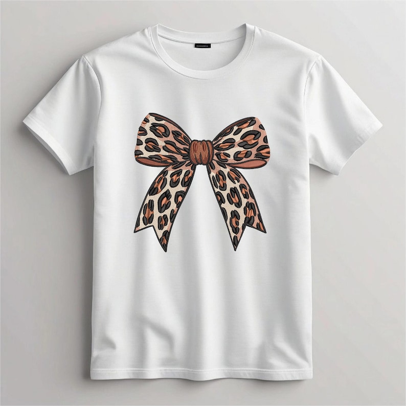 May include: White t-shirt featuring a large, detailed bow design. The bow has a leopard print pattern in shades of brown and tan. The shirt is a classic crew neck style.
