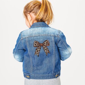 May include: A denim jacket with a faded blue wash. The back of the jacket features a large bow with a leopard print design. The bow has a brown center and tan and brown spots.