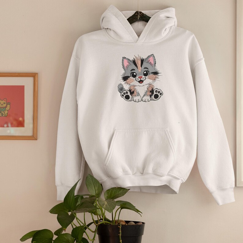 May include: White hooded sweatshirt featuring a colorful embroidered cartoon cat. The cat has gray, pink, and peach fur, with black paws and a smiling expression. The sweatshirt has a front pocket and a drawstring hood.
